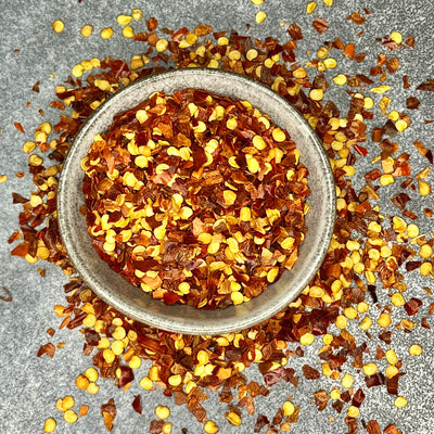 Bright red chilli flakes scattered on a grey surface and in a bowl, showing the coarse texture and intense color of dried, crushed chili peppers, used for adding spice and heat to dishes.