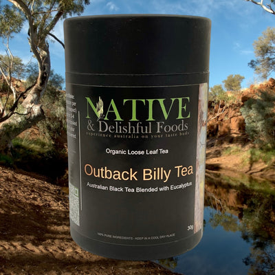 A black canister labeled "Outback Billy Tea" sits prominently in the foreground, with a serene backdrop of a meandering creek surrounded by lush trees. The canister’s design features traditional Australian outback imagery, evoking a sense of rustic charm and natural wilderness. The peaceful creek in the background flows gently through a landscape dotted with various shades of green, reflecting the tranquility and beauty of the Australian bush.